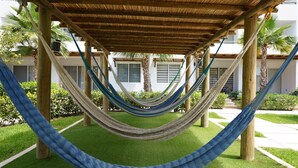 A hammock under a wooden pergola, surrounded by greenery and a building with windows. - Almarena (Mazatlán)