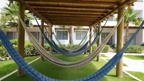 A hammock under a wooden pergola, surrounded by greenery and a building with windows.
