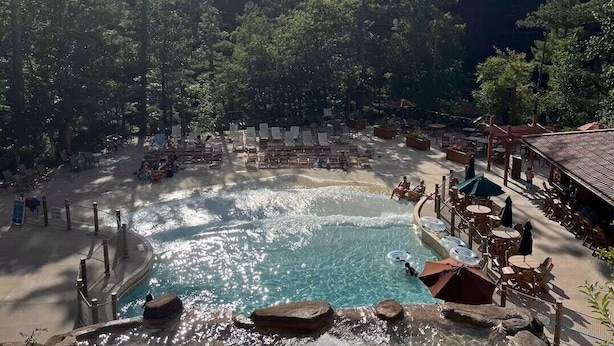 A pool area with lounge chairs and umbrellas, surrounded by trees. - River View Condo - Top Floor - Resort Pools - Pet Friendly! (Wisconsin Dells)