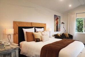 Ein Schlafzimmer mit einem Holzkopfteil, einem großen Bett mit vielen Kissen, Nachttischlampen, einem kleinen Tisch mit Lampe, einem Sessel und einem Bild an der Wand.