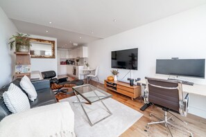 Ein modernes Wohnzimmer mit Sofa, Couchtisch, Fernseher und einem Essbereich, im Hintergrund eine Küche.