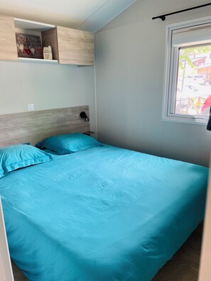 A bedroom with a bed, a nightstand, a wall-mounted shelf with decorative items, and a window with a view of the outside. - 4-person mobile home in a family campsite in the mountains  (Saillagouse)