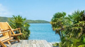 Une terrasse en bois avec une chaise, donnant sur un lac paisible bordé de palmiers et avec des collines en arrière-plan.