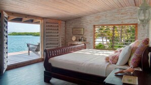 Un plafond en bois, un lit avec des oreillers, une table de chevet, une fenêtre donnant sur l’eau et une terrasse en bois à l’extérieur.