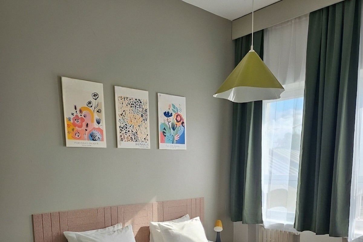 Cama doble con ropa blanca, cabecero y mesita de noche con lámpara. Tres obras de arte decoran la pared sobre la cama.