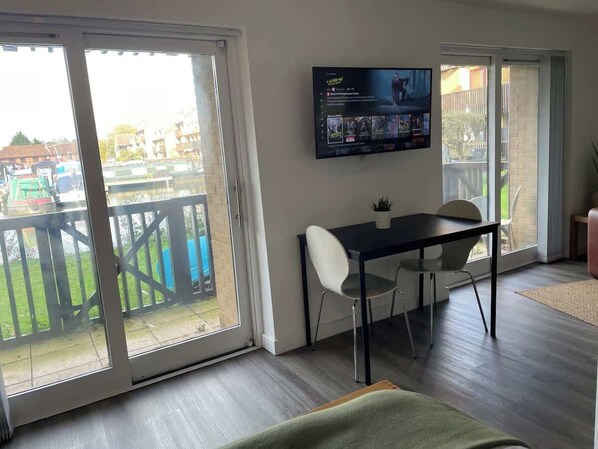A room with a sliding glass door leading to a balcony, a flat-screen TV mounted on the wall, a dining table with chairs, and a small potted plant.