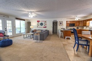 A spacious living area with a grey sofa, a coffee table, and a blue bean bag. A kitchen with wooden cabinets and a dining area with a wooden table and chairs is visible.