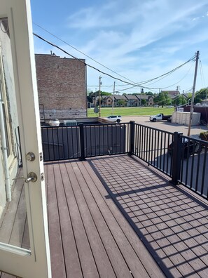 A wooden deck with a black railing, a brick wall, and a clear view of a street with parked cars and houses in the background.