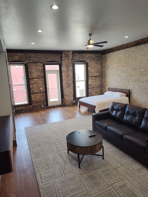 A spacious room with a bed, a black leather sofa, a round coffee table, and a ceiling fan.