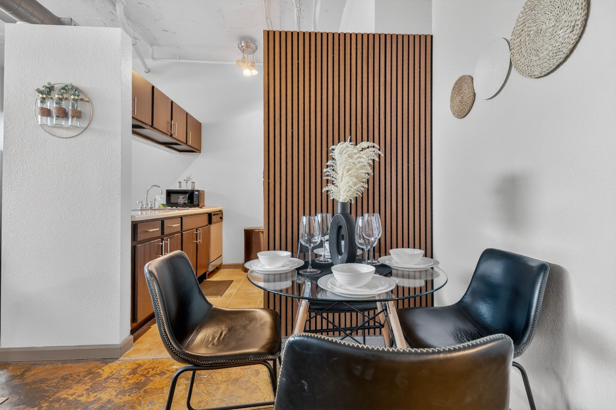 Un espace repas comprenant une table ronde en verre, des chaises en cuir noir et une cloison en bois avec des lattes verticales.