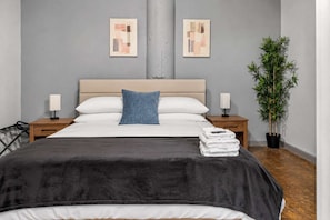 Ein modernes Hotelzimmer mit einem großen Bett, zwei Nachttischen mit Lampen, einem blauen Kissen, einem Stapel weißer Handtücher und einer Topfpflanze.