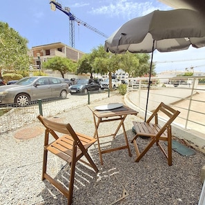 An outdoor seating area with wooden chairs and a table, a covered umbrella, and a view of parked cars and buildings in the background. - 1-Bedroom Apartment Near the Beach in Cabanas with a Parking Space (Cabanas de Tavira)
