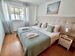 A hotel room with two beds, a window with curtains, a bedside table with a lamp, and a painting on the wall. - 1-Bedroom Apartment Near the Beach in Cabanas with a Parking Space (Cabanas de Tavira)