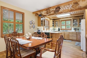 A dining area with a wooden table and chairs, a kitchen with wooden cabinets and a refrigerator, and a window with blinds. - Lakefront Lodge w Dock, Fire Pit & Views! (Cullowhee)