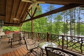 A covered outdoor deck with wooden flooring, metal chairs, a glass table, and a railing overlooking a forested area. - Lakefront Lodge w Dock, Fire Pit & Views! (Cullowhee)