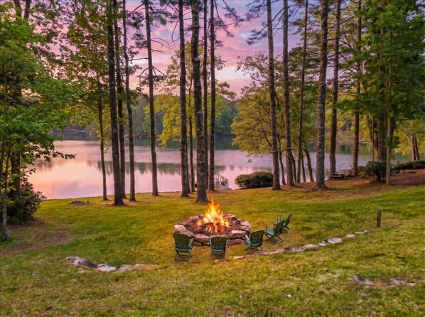 A lakeside campfire with chairs, surrounded by tall trees and a calm lake. - Lakefront Lodge w Dock, Fire Pit & Views! (Cullowhee)