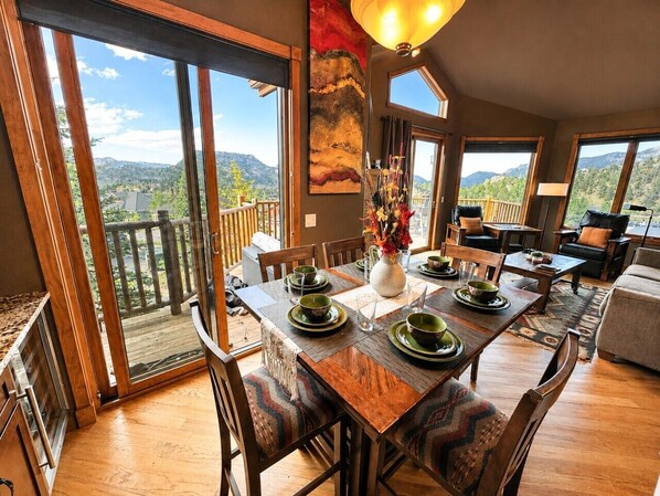 Un comedor con una mesa de madera para cuatro personas, ventanas amplias con vista a la montaña y un balcón con barandilla de madera.