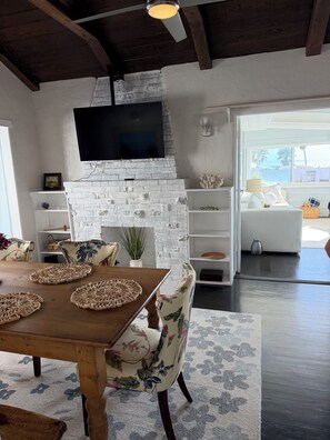 Een woonkamer met een witte bakstenen open haard, een flatscreen televisie erboven, een houten eettafel met geweven placemats, stoelen met bloemenprint en uitzicht op de oceaan door een groot raam.