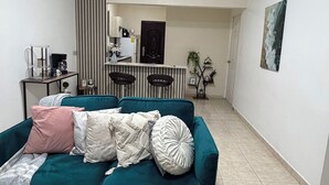 Una sala de estar con un sofá verde azulado, una mesa de centro y una cocina al fondo.