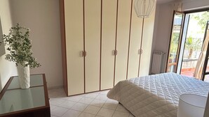 Ein Schlafzimmer mit Bett, Kleiderschrank, Nachttisch, einer Pendelleuchte und Blick durch ein Fenster.