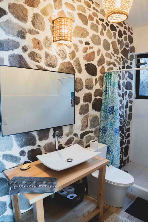 Un baño con pared de piedra, un lavabo blanco, un espejo, una toalla y una cortina de ducha.