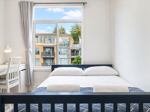 Ein Schlafzimmer mit Bett, Schreibtisch und Stuhl, ein Fenster mit Blick auf Gebäude und einer Lampe.