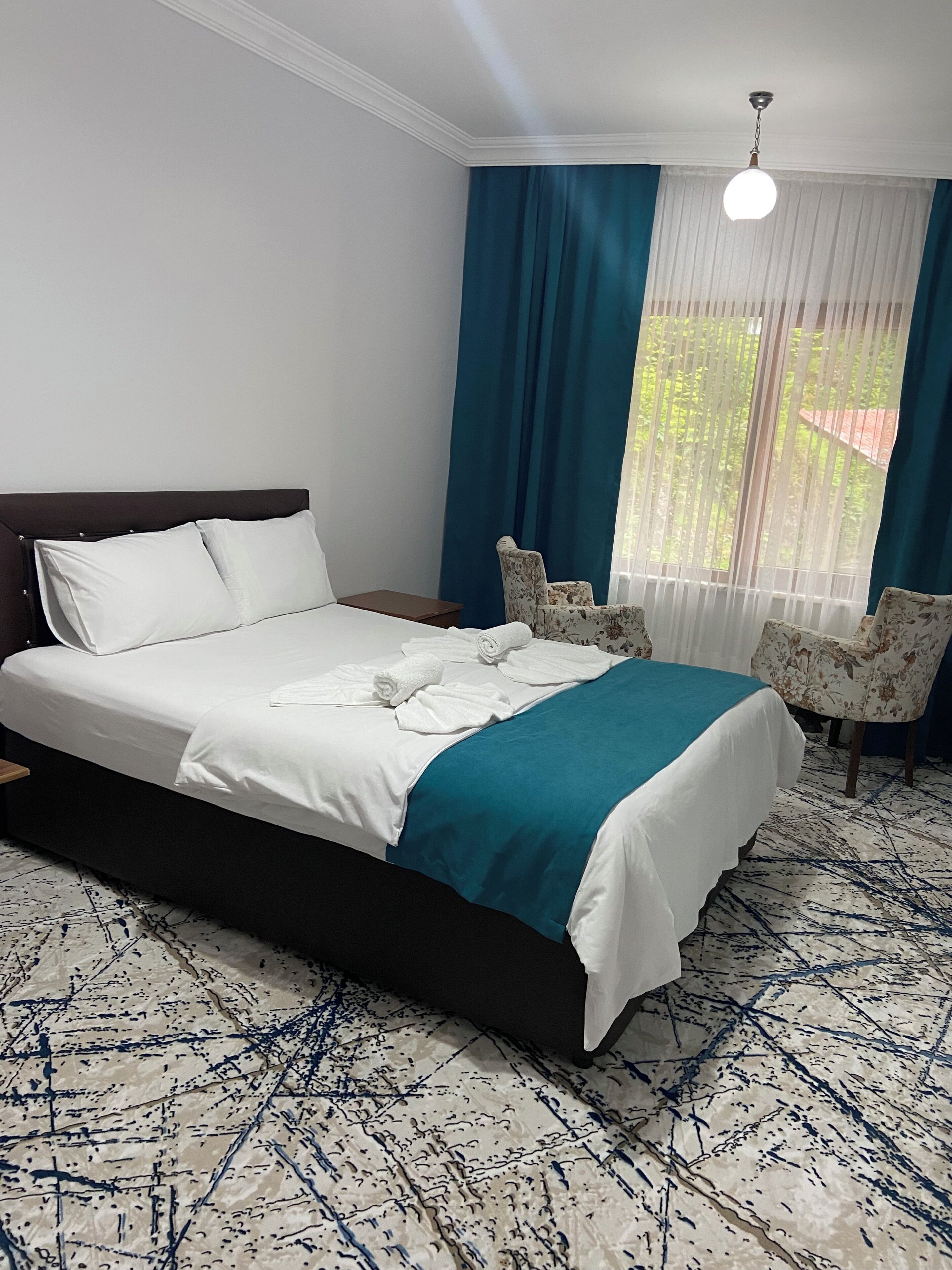 Una cama bien hecha con sábanas blancas y una manta azul turquesa, una mesita de noche de madera, dos sillas con estampado floral, un ventanal con cortinas azules y un suelo estampado.
