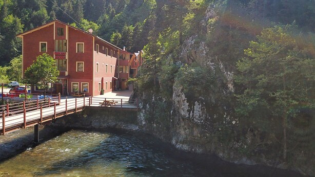 A red building with a bridge over a river, surrounded by trees. - soylu otel restoran (Caykara)