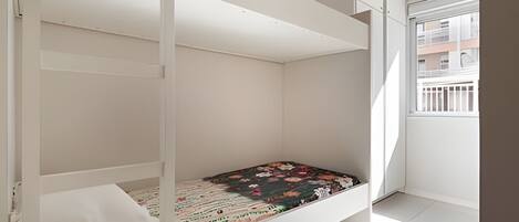 Una cama litera con colchón estampado con motivos florales, paredes blancas y una ventana con vistas a edificios.