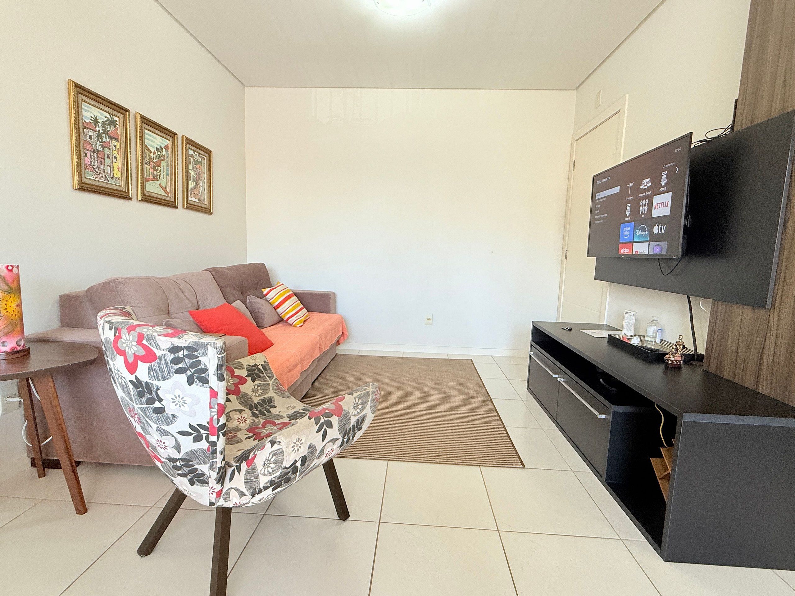 Een woonkamer met een bank, een bloemen fauteuil, een televisie en lijstjes met foto's aan de muur.