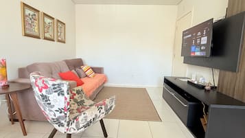 Een woonkamer met een bank, een bloemen fauteuil, een televisie en lijstjes met foto's aan de muur.
