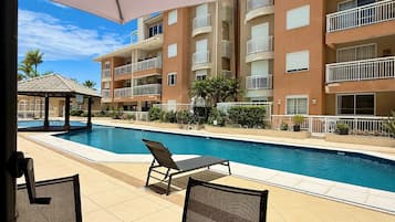 A swimming pool with lounge chairs, a covered area, and a multi-story building with balconies.