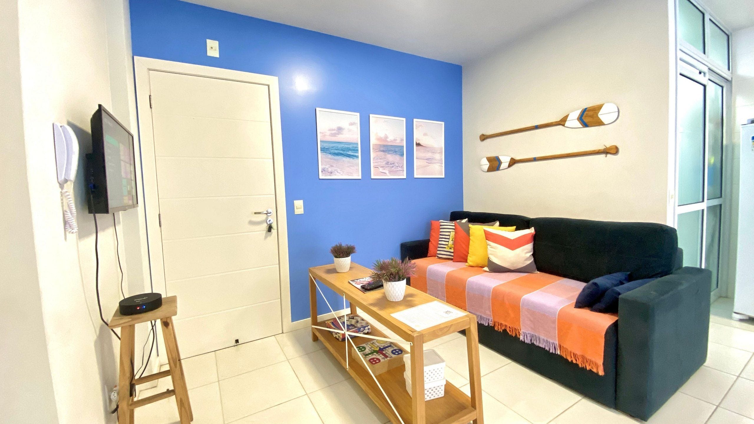 A room with a blue wall, a wooden door, a black TV, a wooden table with a remote, a green sofa with colorful cushions, and three framed pictures.