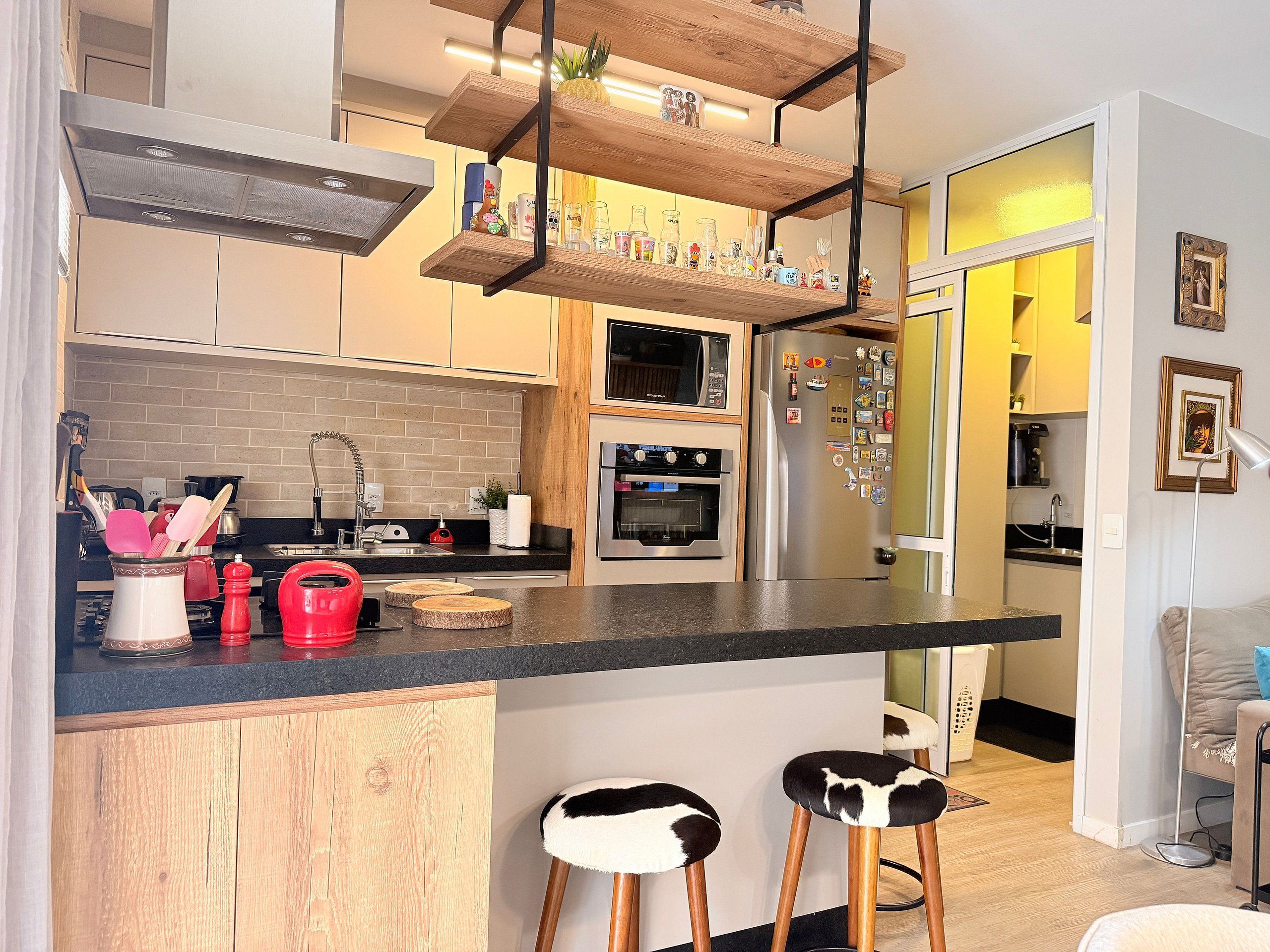 Eine moderne Küche mit Einbauofen, Mikrowelle und Kühlschrank, Holzbarhocker und einem hängenden Regal mit verschiedenen Gegenständen.