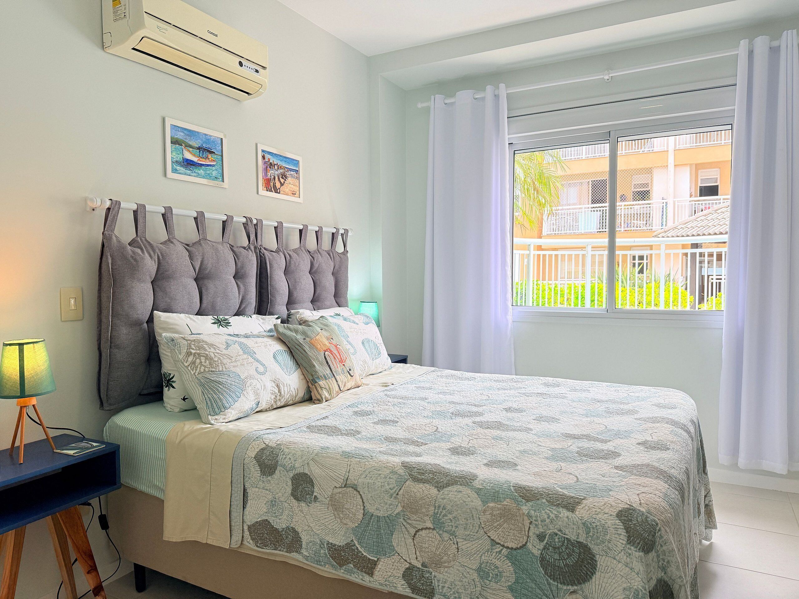 A bedroom with a bed, air conditioning unit, two paintings, a window with curtains, and a lamp.