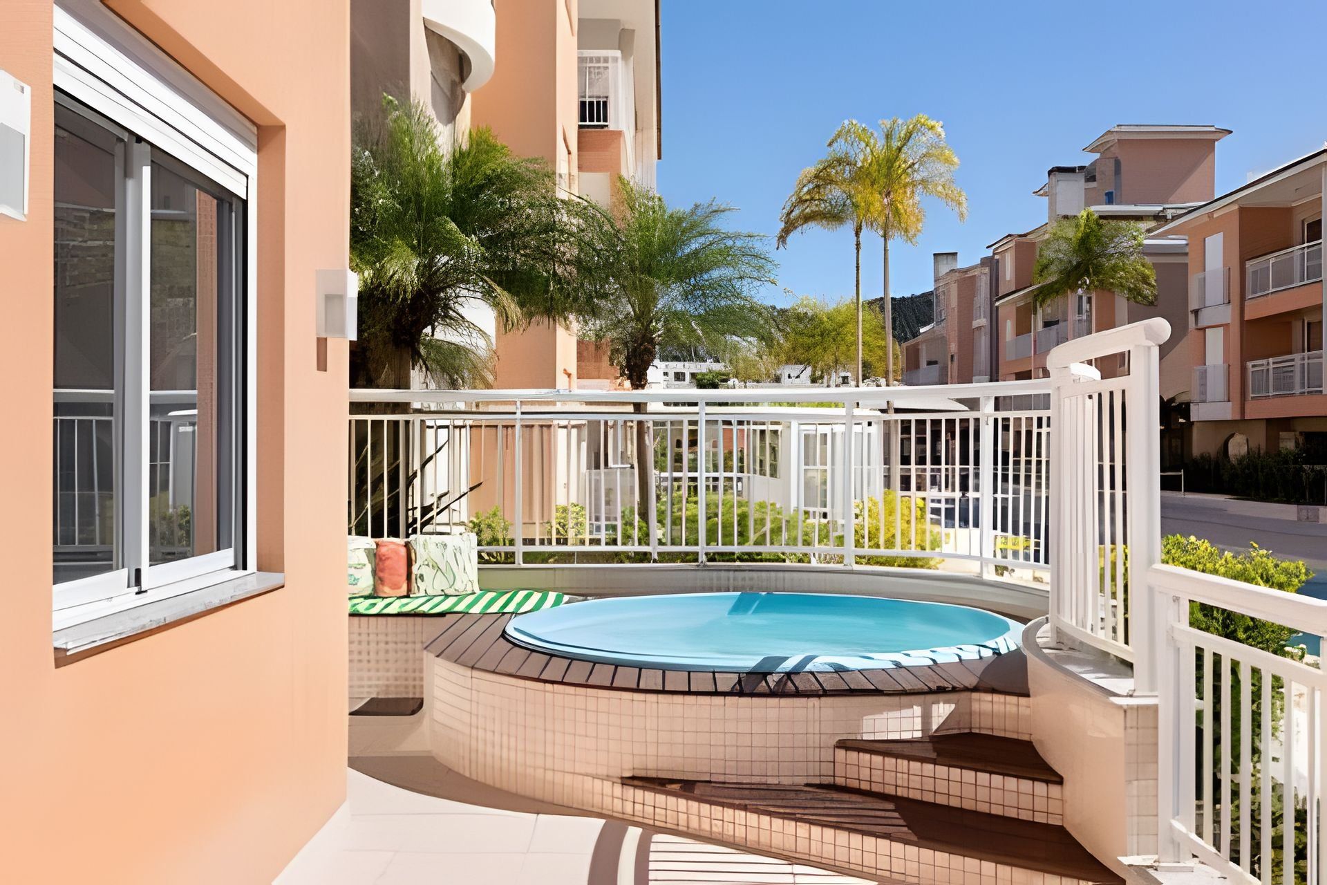 Un edificio residencial con piscina, un balcón con baranda blanca y vistas a edificios vecinos y palmeras.