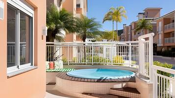 A residential building with a swimming pool, a balcony with a white railing, and a view of neighboring buildings and palm trees.