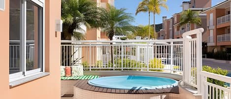 A residential building with a swimming pool, a balcony with a white railing, and a view of neighboring buildings and palm trees.