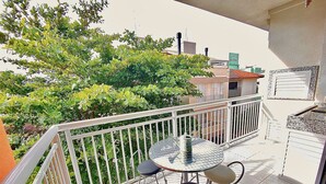 A balcony with a round table and two chairs, surrounded by a white railing, overlooking a green area with trees and buildings in the background.