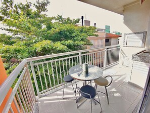 A balcony with a round table and two chairs, surrounded by a white railing, overlooking a green area with trees and buildings in the background. - Residencial com piscina (Florianópolis)
