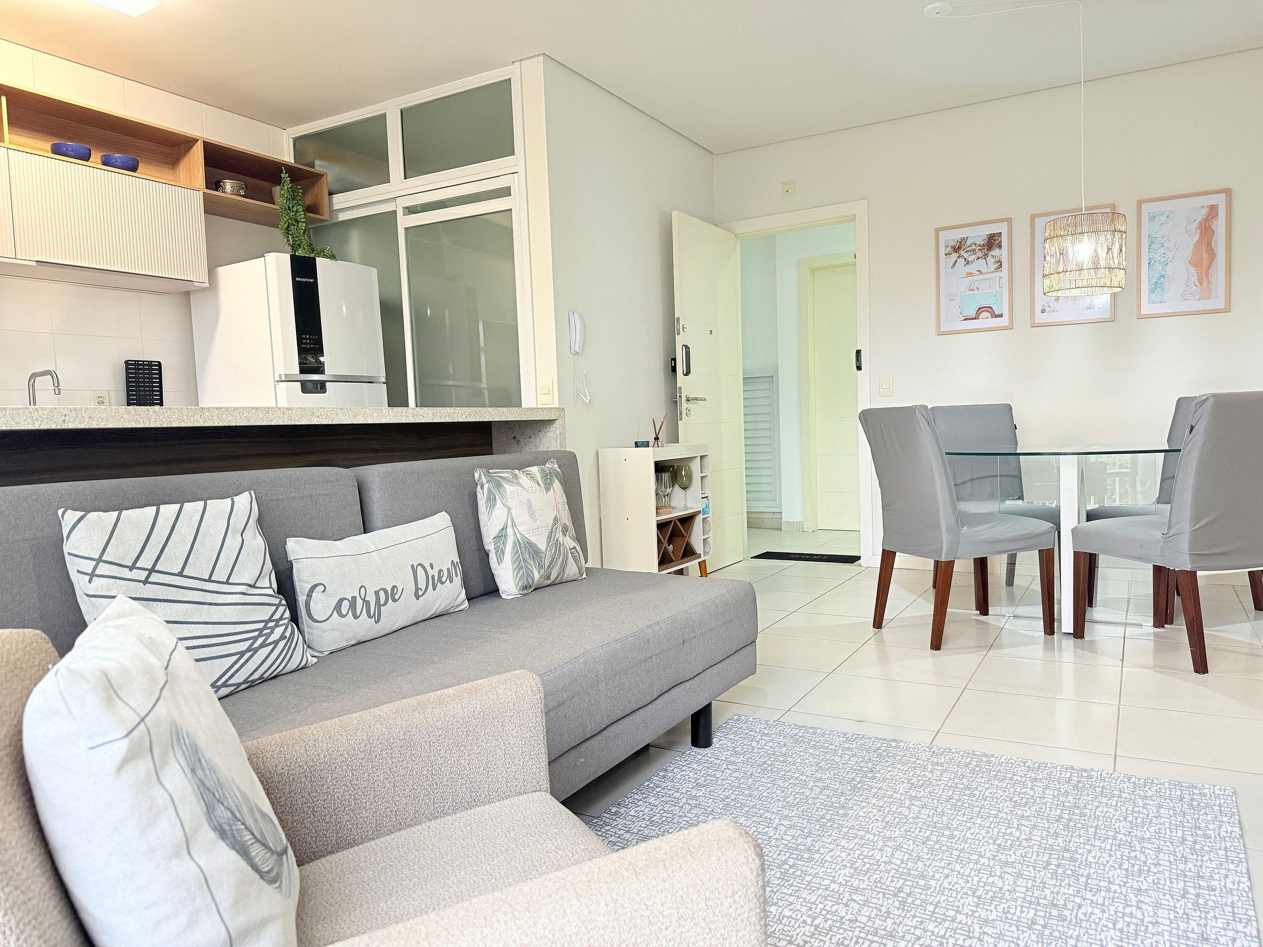A modern living room with a grey sofa, a dining table with chairs, and a kitchen area with white cabinets and a plant.