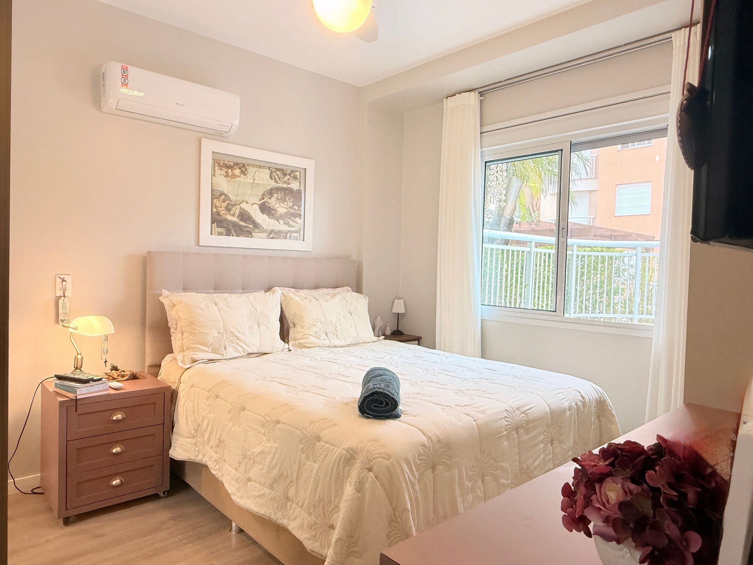 A bedroom with a bed, bedside tables, a nightstand, a wall-mounted air conditioner, a framed picture, and a window with a view of the outside.
