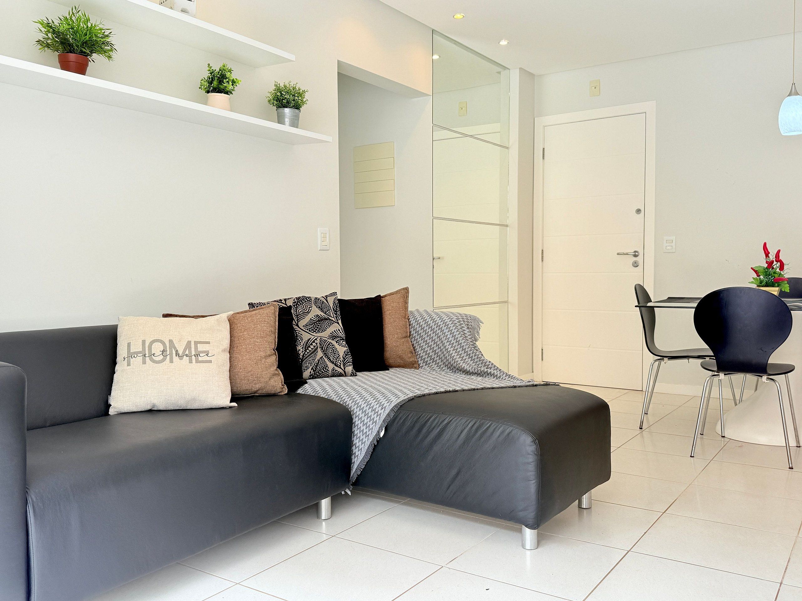 A modern living room with a grey sofa, a small table, and a chair. There are potted plants on a shelf above the sofa.