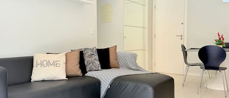 A modern living room with a grey sofa, a small table, and a chair. There are potted plants on a shelf above the sofa.
