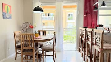 A dining area with a round table, wooden chairs, a fan, and a view of a balcony with red curtains.