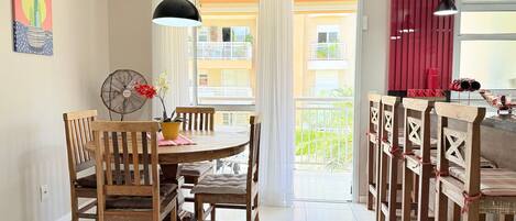 A dining area with a round table, wooden chairs, a fan, and a view of a balcony with red curtains.