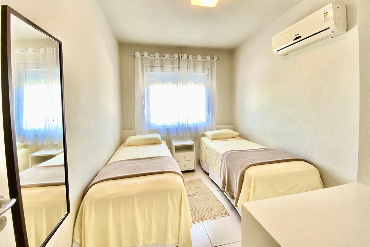 A hotel room with two single beds, a desk, a mirror, a window with sheer curtains, and an air conditioning unit on the wall.