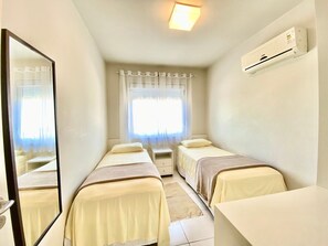 A hotel room with two single beds, a desk, a mirror, a window with sheer curtains, and an air conditioning unit on the wall. - Residencial com piscina (Florianópolis)