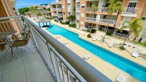 A hotel pool area with a clear view of the pool, lounge chairs, and a building with balconies.