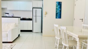 A modern kitchen with white cabinets, a refrigerator, and a dining area with a white table and chairs. - Residencial com piscina (Florianópolis)